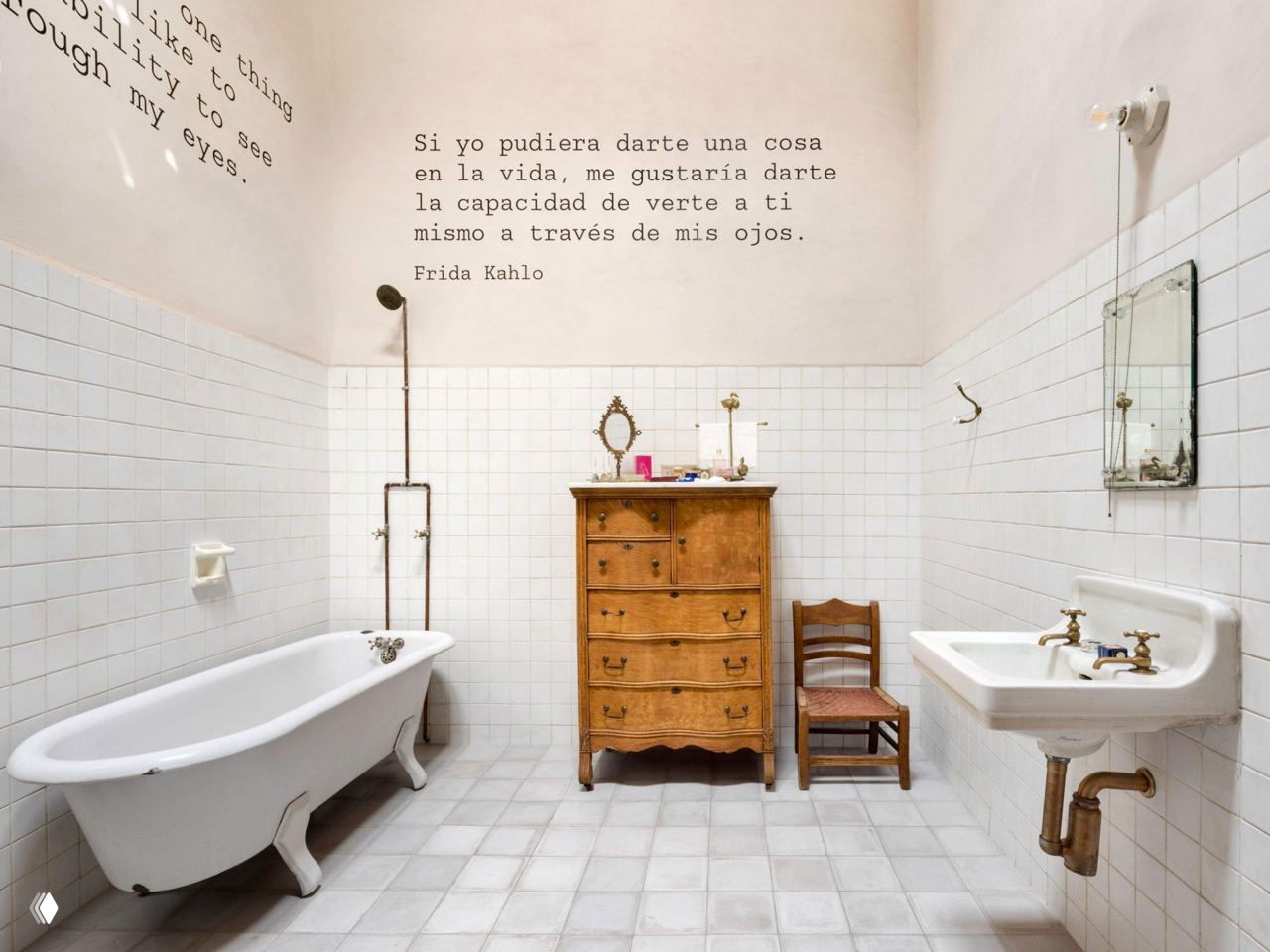 Ванная комната с чугунной ванной, деревянным комодом и кафельной облицовкой; на белой стене видна цитата Фриды Кало, интерьер освещён естественным светом.