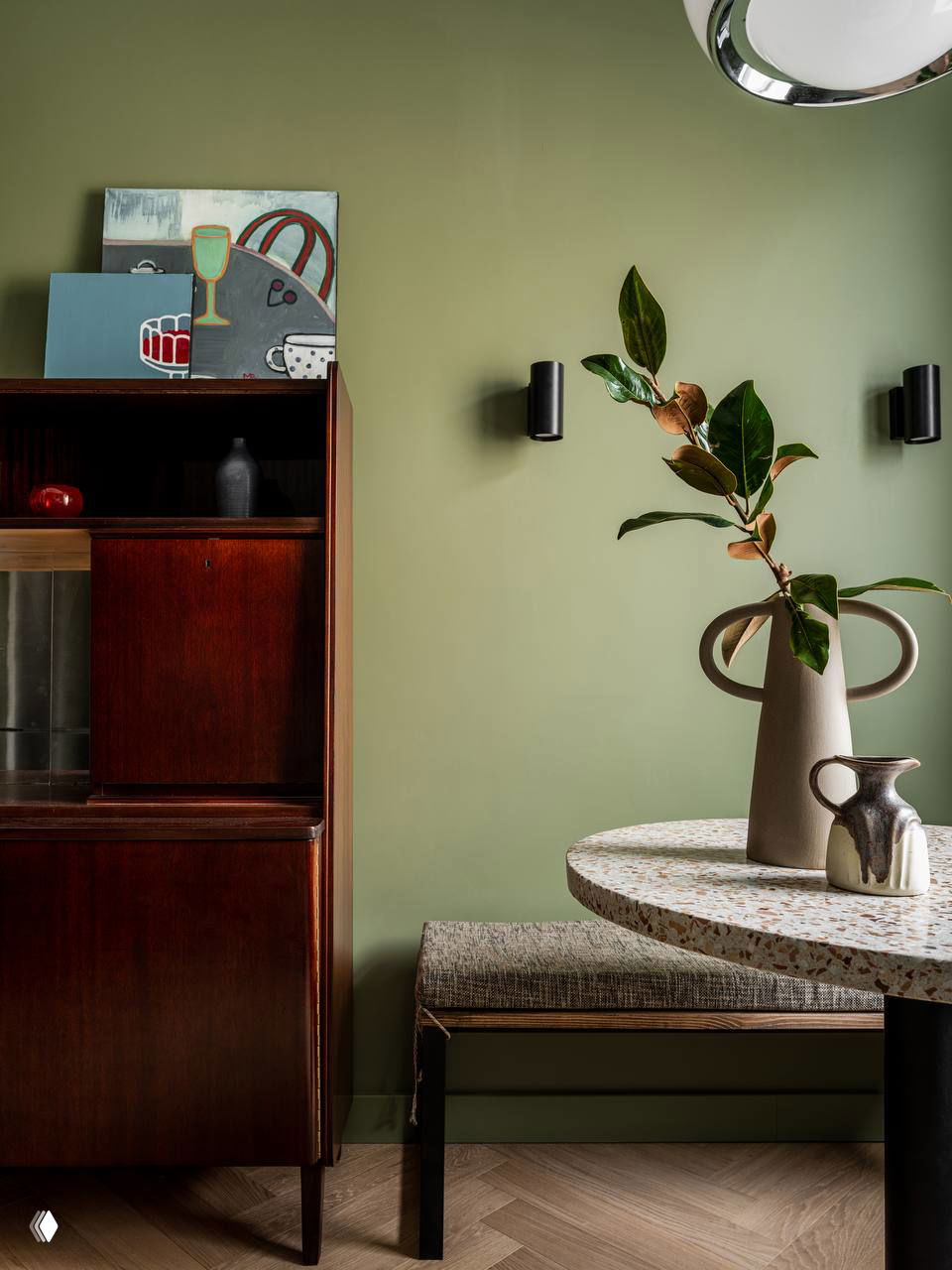 Интерьерный угол с оливковыми стенами, тёмный деревянный буфет, круглый стол и большая ваза с глянцевыми зелёными листьями на переднем плане.