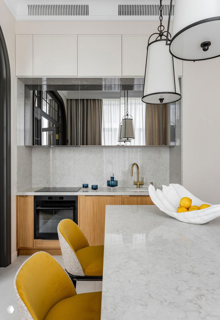 Мини-кухня с каменной столешницей и барной стойкой, барные стулья с жёлтой обивкой и золотистый смеситель — контраст тёплой древесины и светлого камня.