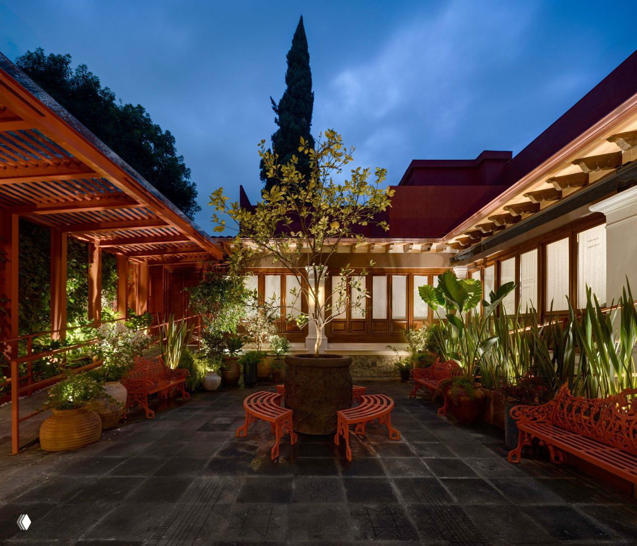 Внутренний двор Museo Casa Kahlo вечером: подсвеченные растения, красные скамьи и деревянные элементы вокруг центрального круглого фонтана, атмосфера сада в центре дома.