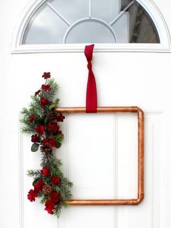 Дверная идея: медная рамка с привязанной красной лентой и веткой ели с ягодами и несколькими шишками в качестве минималистичного праздничного декора.