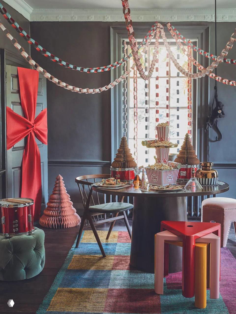 Комната с гирляндами и бумажными украшениями над столом: центральный стол сервирован в тёплых тонах, рядом декоративные ёлочки и подарки.