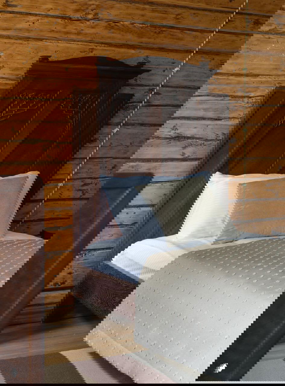 Деталь интерьера: резное деревянное изголовье кровати на фоне бревенчатой стены, текстура дерева и спокойные оттенки постельного белья.