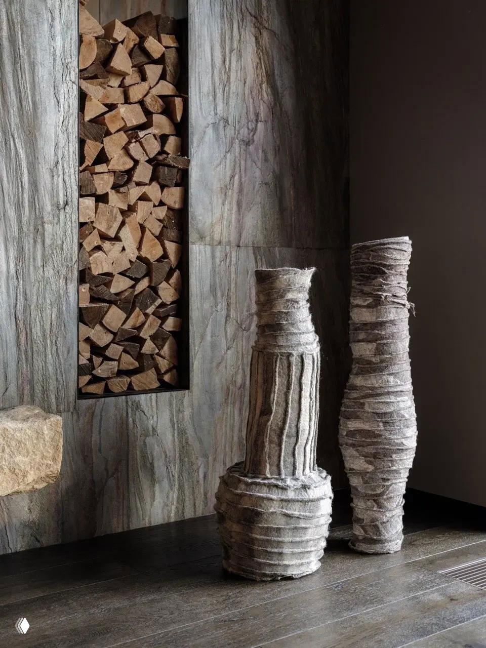 Две высокие текстильные вазы из серого войлока с слоистой структурой стоят рядом с нишей для дров, подчёркивая натуральную фактуру материала.