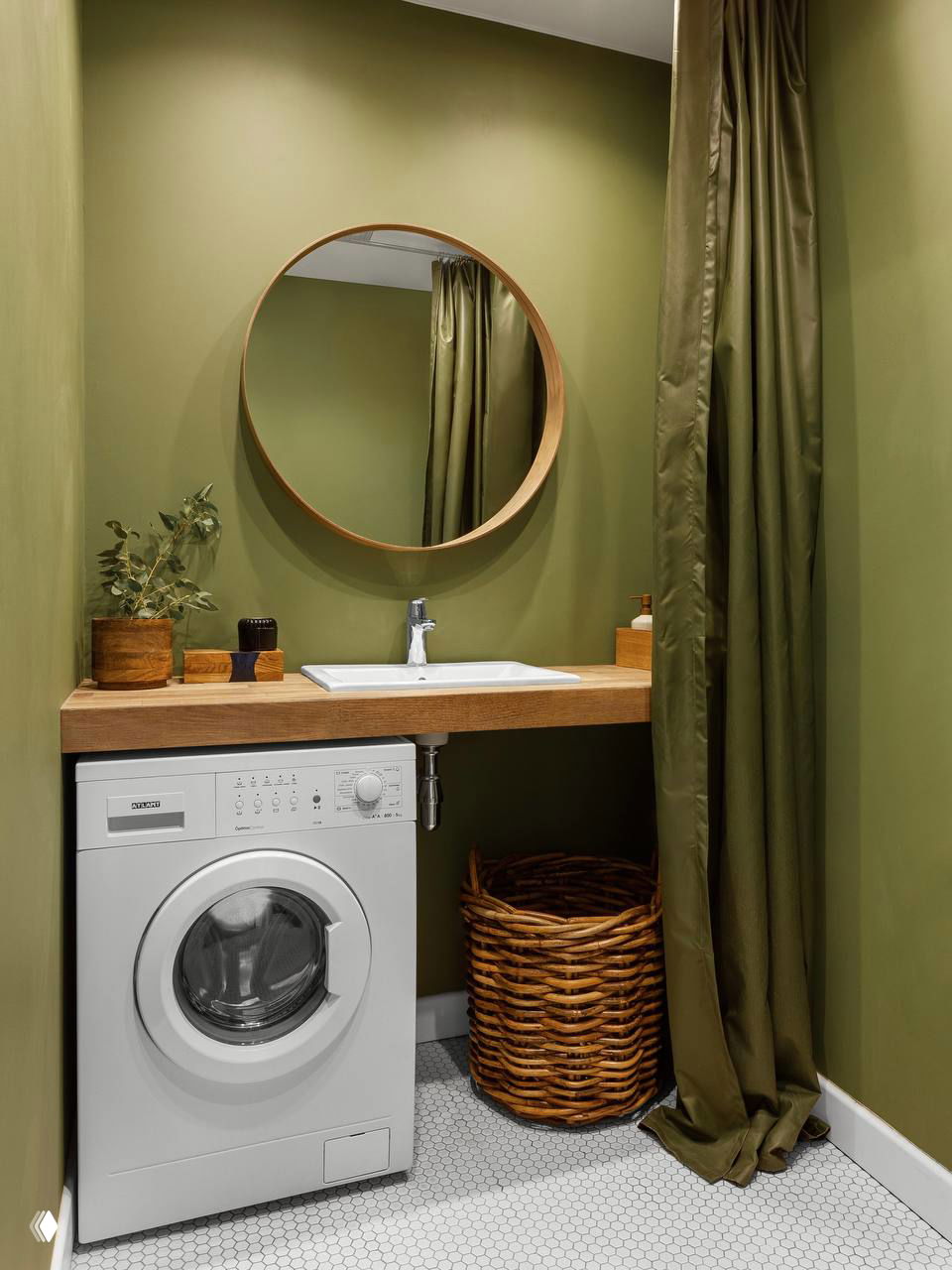 Прачечная-ниша с оливковой окраской стен, встроенной стиральной машиной под деревянной столешницей и большой плетёной корзиной для белья у пола.
