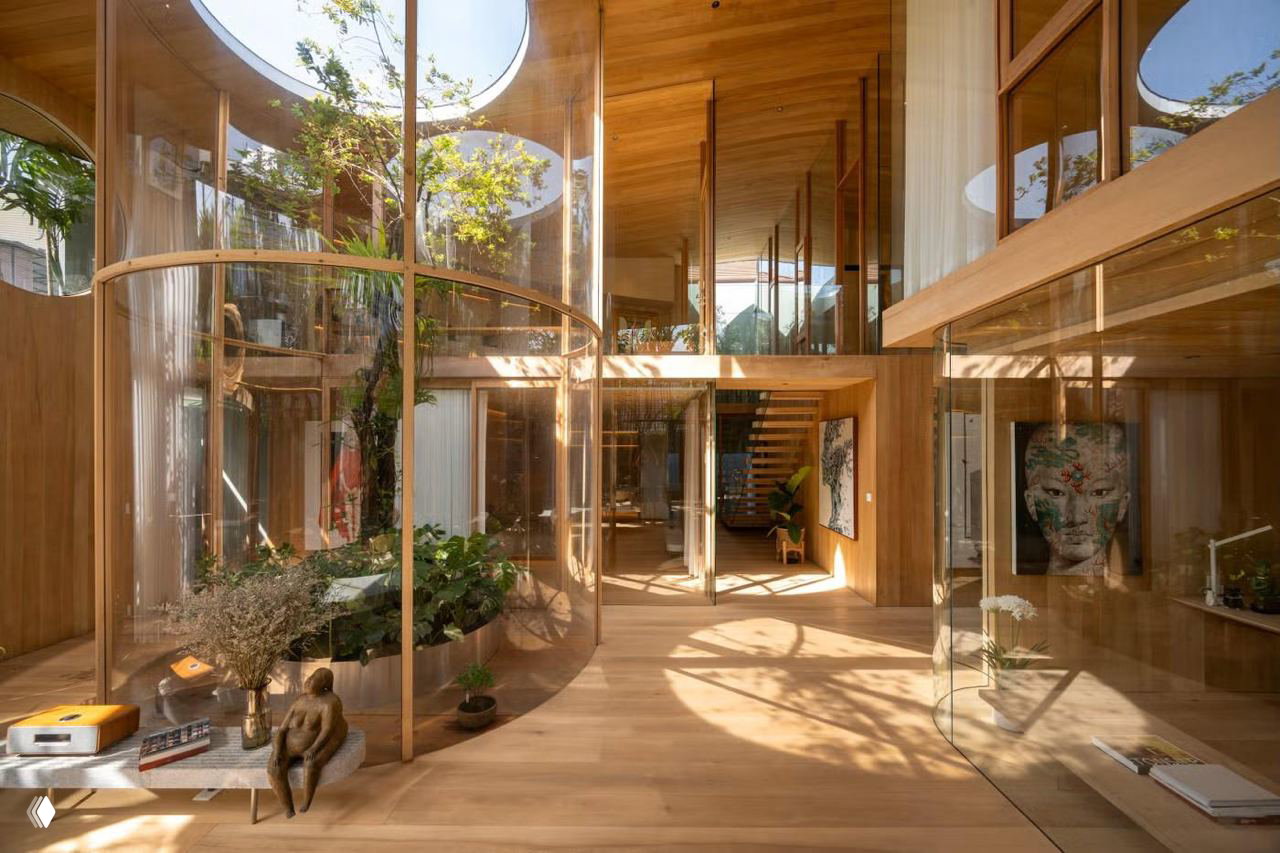 Интерьер дома-оранжереи в Таиланде: стеклянные колонны и круглые световые проёмы вокруг взрослых деревьев, тёплый деревянный пол и графика движущихся теней.