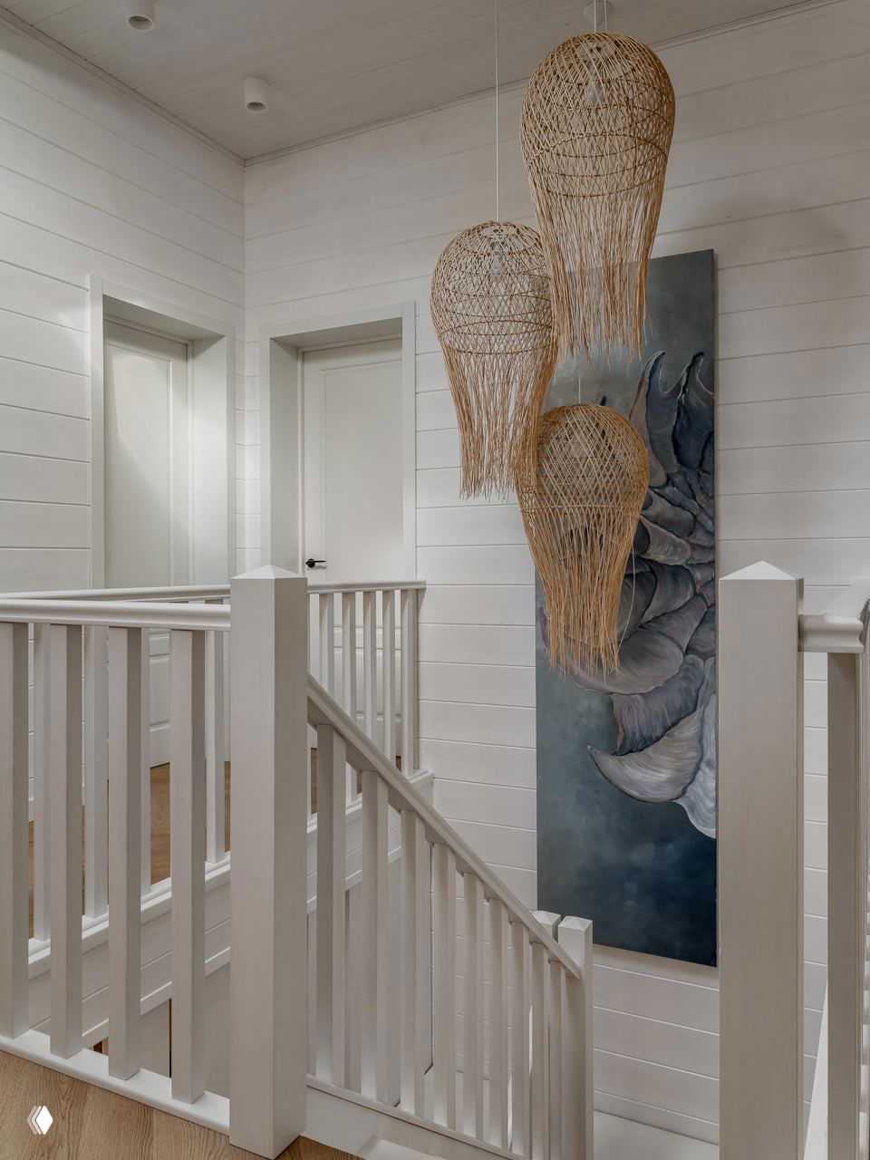 Лестничная зона с белой деревянной перилой и группой ажурных плетеных светильников, крупная настенная картина на фоне светлого убранства.