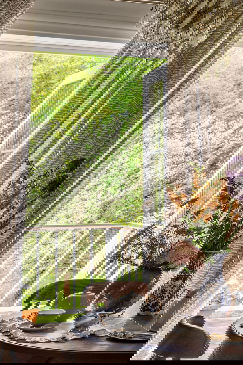 Вид через открытые двери: стол у окна с вазами и сервировкой, перила и зелёный сад за домом создают дачную атмосферу.