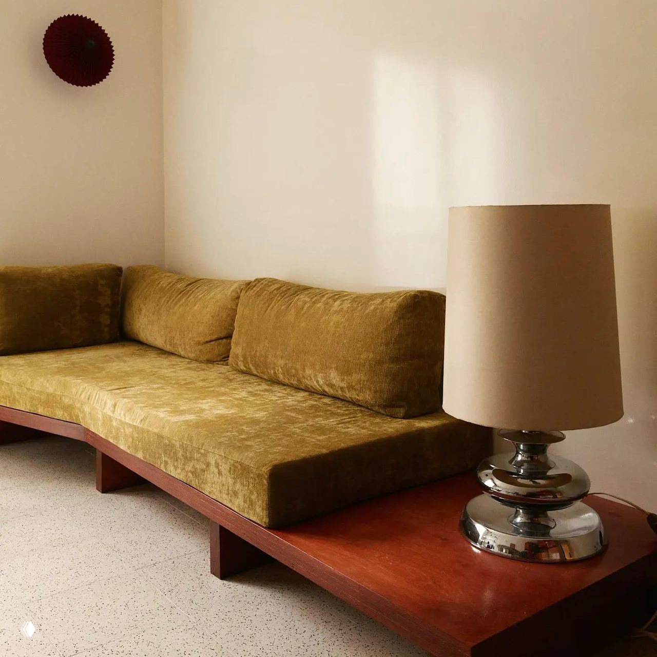 Угловой диван оливково-охрового цвета на низкой деревянной платформе, металлическая лампа и минималистичная стена в светлых тонах.