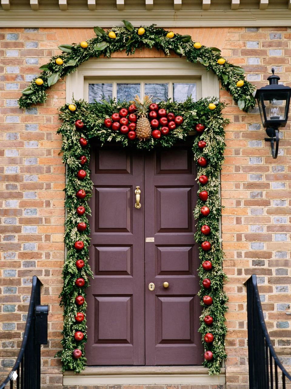 Коричневая дверь в кирпичном фасаде обрамлена гирляндой с красными декоративными яблоками и ананасом сверху — нетипичный натуральный вариант рождественского декора.