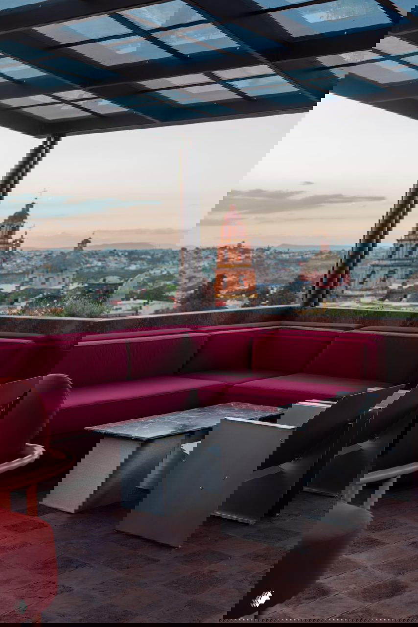 Крыша‑бар под стеклянной навесной конструкцией: угловой бордовый диван, металлические кресла и панорама исторического города с куполами на заднем плане.