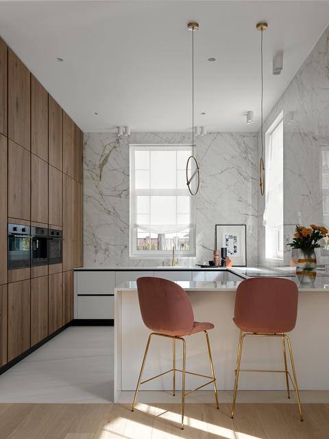 Кухня-столовая в доме Чекалиных: кухонный остров с мраморной стеной, барные стулья розового оттенка и тёплые деревянные шкафы.