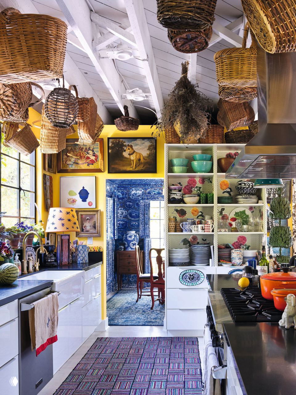 Кухня модельера с навесными корзинами на потолке, жёлтыми стенами, открытыми полками с посудой и взглядом в комнату с синим текстилем за дверным проёмом.