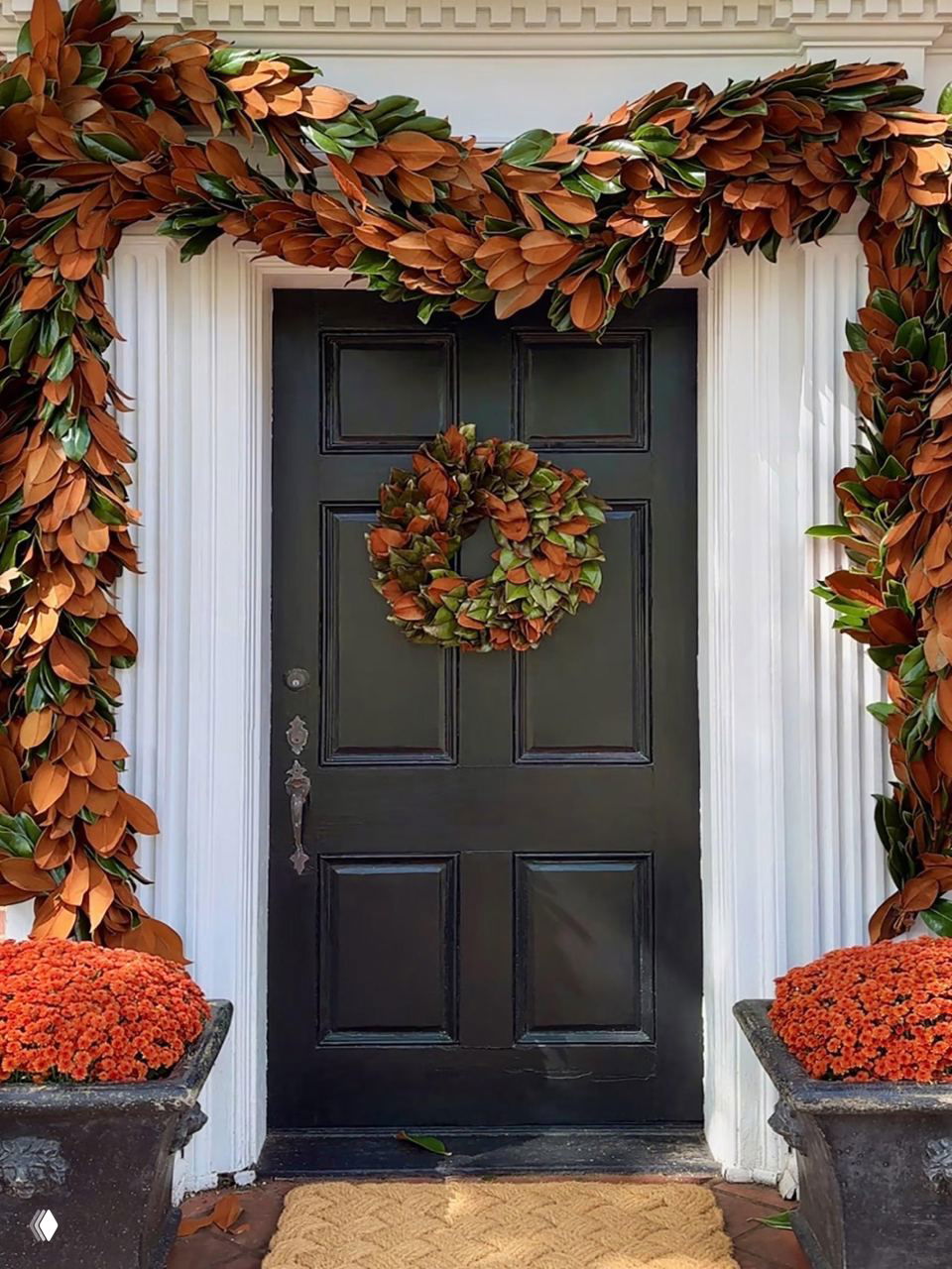 Темная дверь окружена большой гирляндой из коричневых и зелёных листьев магнолии, центральный венок из тех же материалов создаёт спокойную природную палитру декора.
