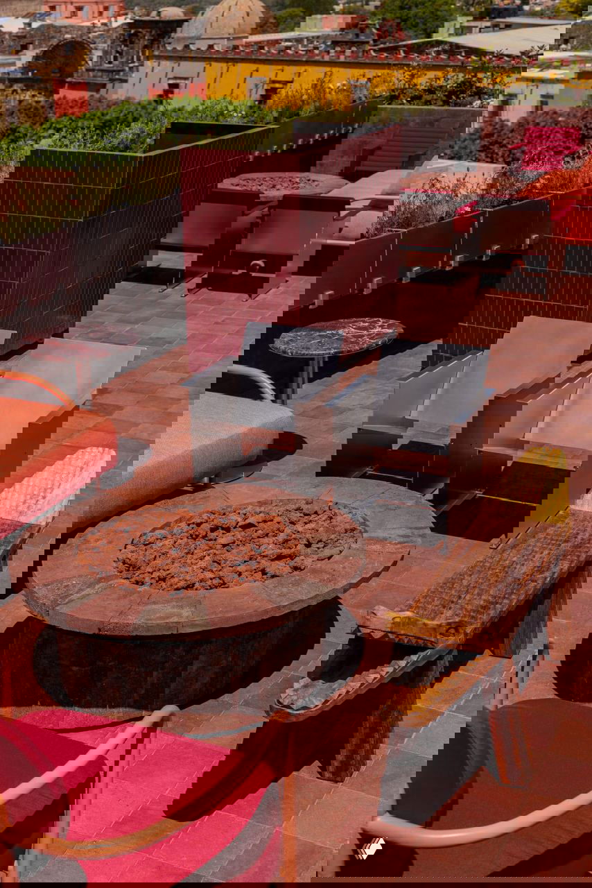 Низкий круглый стол‑очаг, плитка тёмно‑красного цвета и две стальные кресла рядом с деревянными скамьями — сочетание тесонтле и ремесленных элементов.