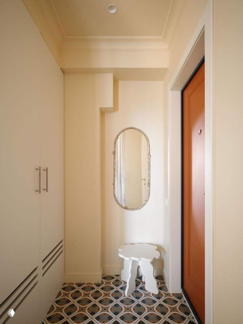 Прихожая с декоративным зеркалом на стене, белым табуретом и тёплой входной дверью терракотового оттенка; узорная напольная плитка и лаконичная мебель.