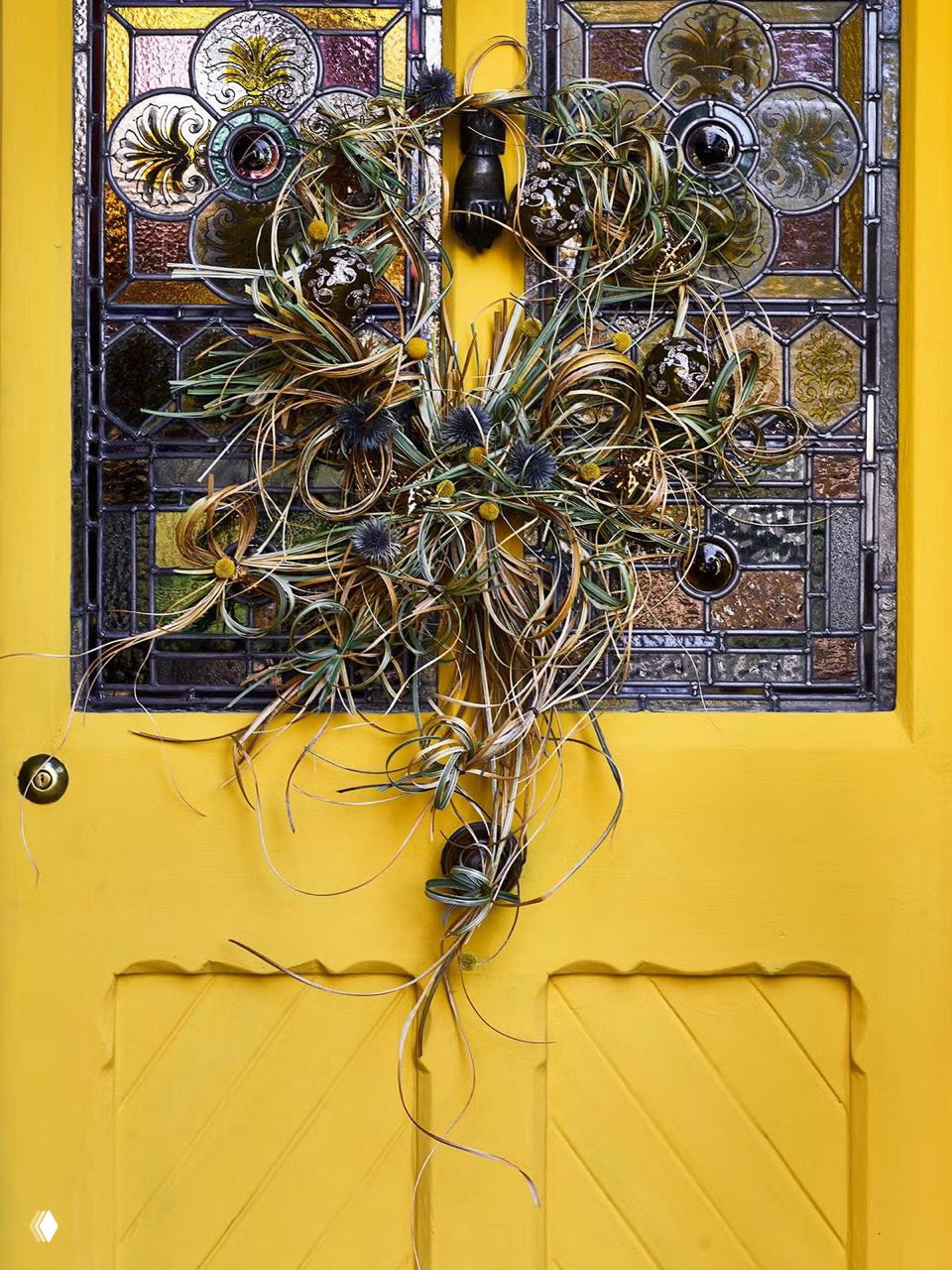 Жёлтая резная дверь украшена декоративным венком из сухих трав и плетений, витражное стекло на двери дополняет текстурный, арт‑ориентированный вариант праздничного декора.