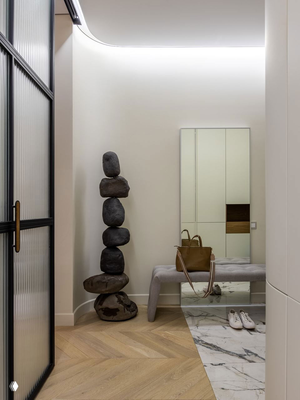 Прихожая с высоким потолком, декоративной инсталляцией из камней в виде столба и скамьей у зеркала — спокойная нейтральная палитра и паркет елочкой.