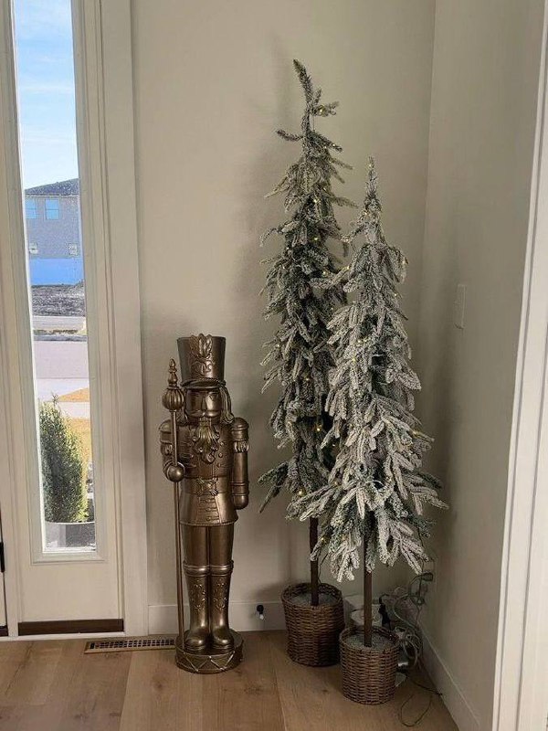 Две заснеженные худые ёлки у входной двери рядом с декоративным щелкунчиком — вертикальная композиция в светлом прихожем с деревянным полом.