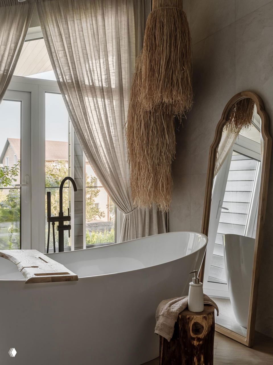 Ванная комната с отдельно стоящей белой ванной, большим зеркалом в деревянной раме и длинными декоративными подвесными элементами из натуральных волокон, вид на природу за окном.