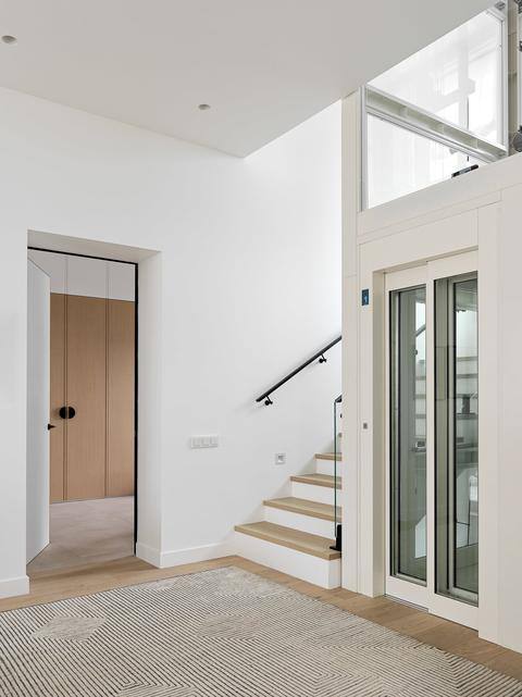 Прихожая и холл дома: белые стены, лестница с деревянными ступенями, компактный лифт и минималистичный интерьер в светлой гамме.