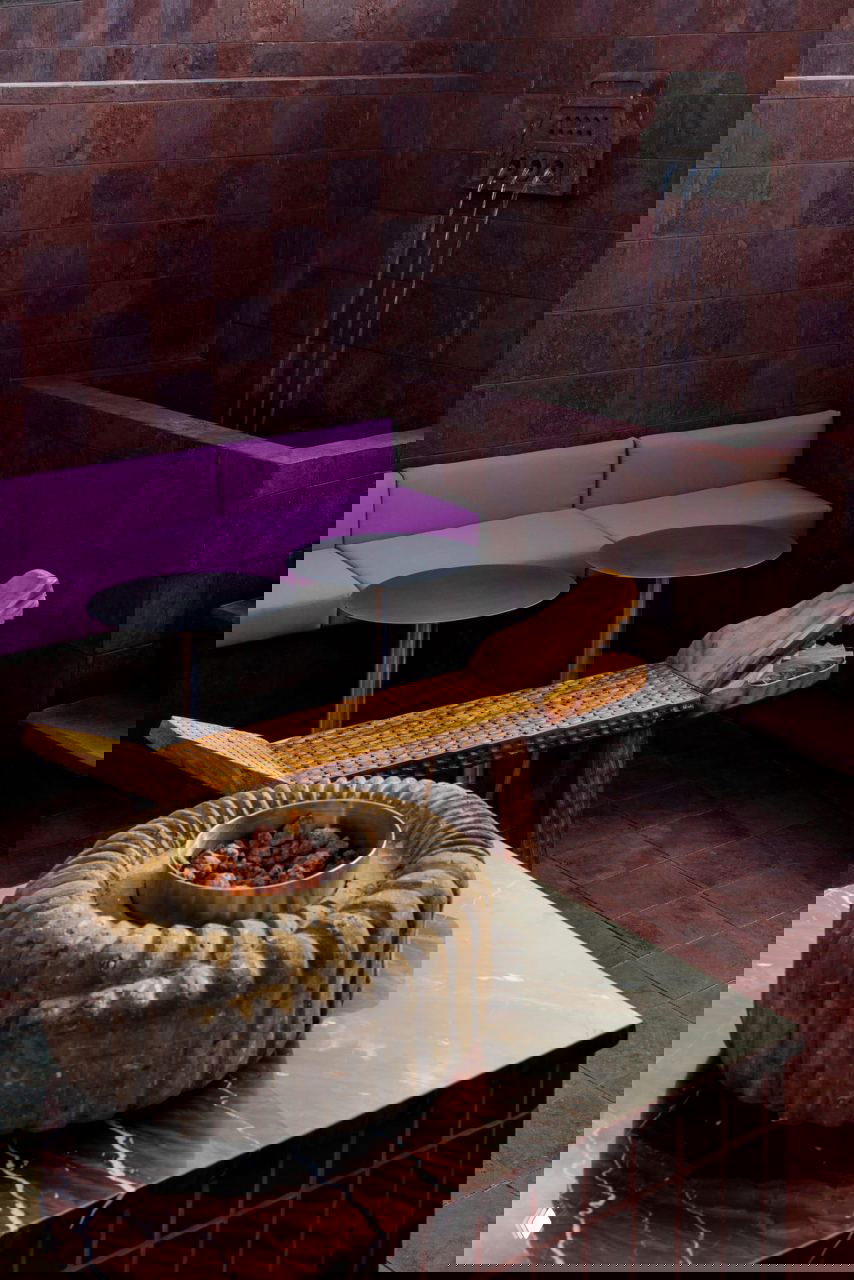 Уютный угол с розово‑фиолетовым диваном, декоративным фонтаном и резной деревянной лавкой на фоне стен, облицованных плиткой из вулканического камня тесонтле.