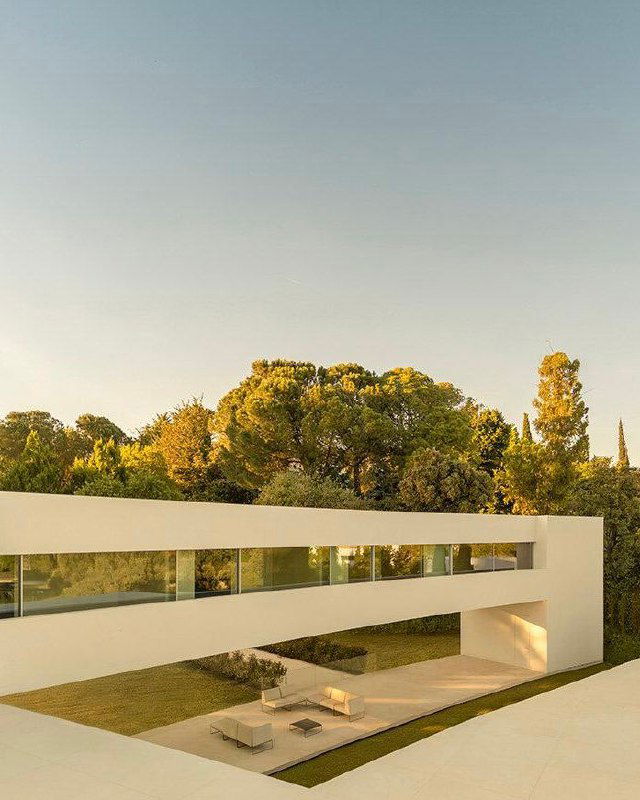 Вид на внутренний дворик Villa Lago: белая вытянутая галерея над открытой зоной с лаунж-площадкой и зелёным газоном, характерная игра света и тени.