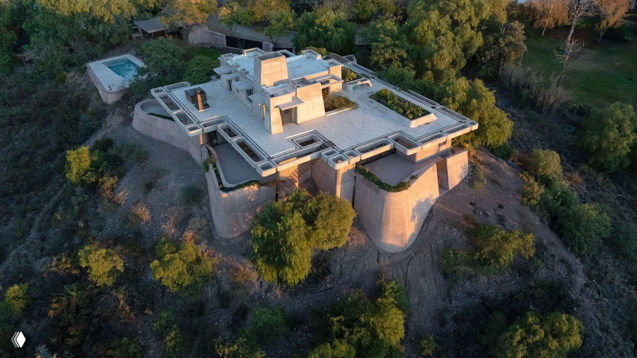 План сверху: плоская кровля и мощный полигональный контур дома, садовые участки и небольшой бассейн на фоне зелёного оазиса внизу.