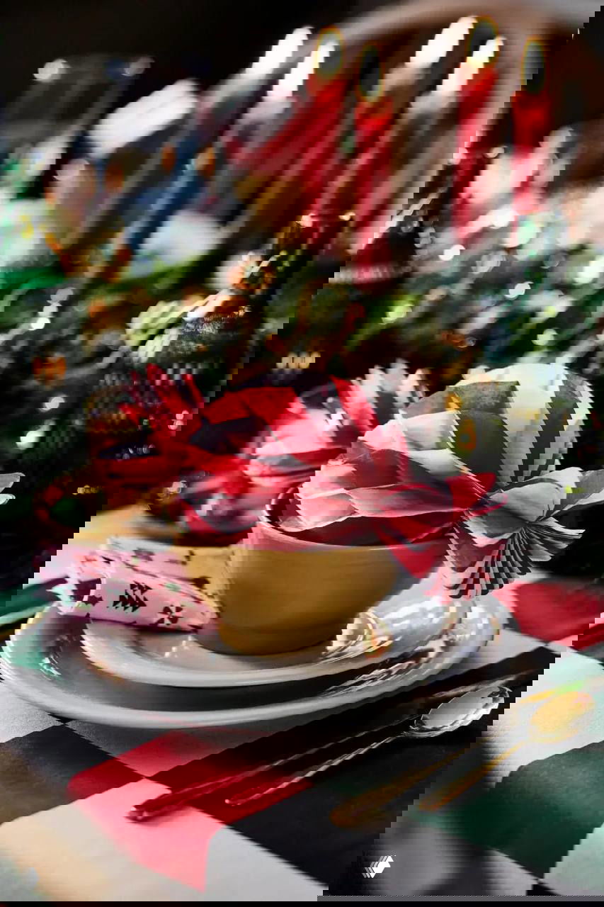 Праздничная сервировка: тарелки, тканевая салфетка и бумажные банты в красно‑белой гамме на фоне свечей и зелёных бутылок