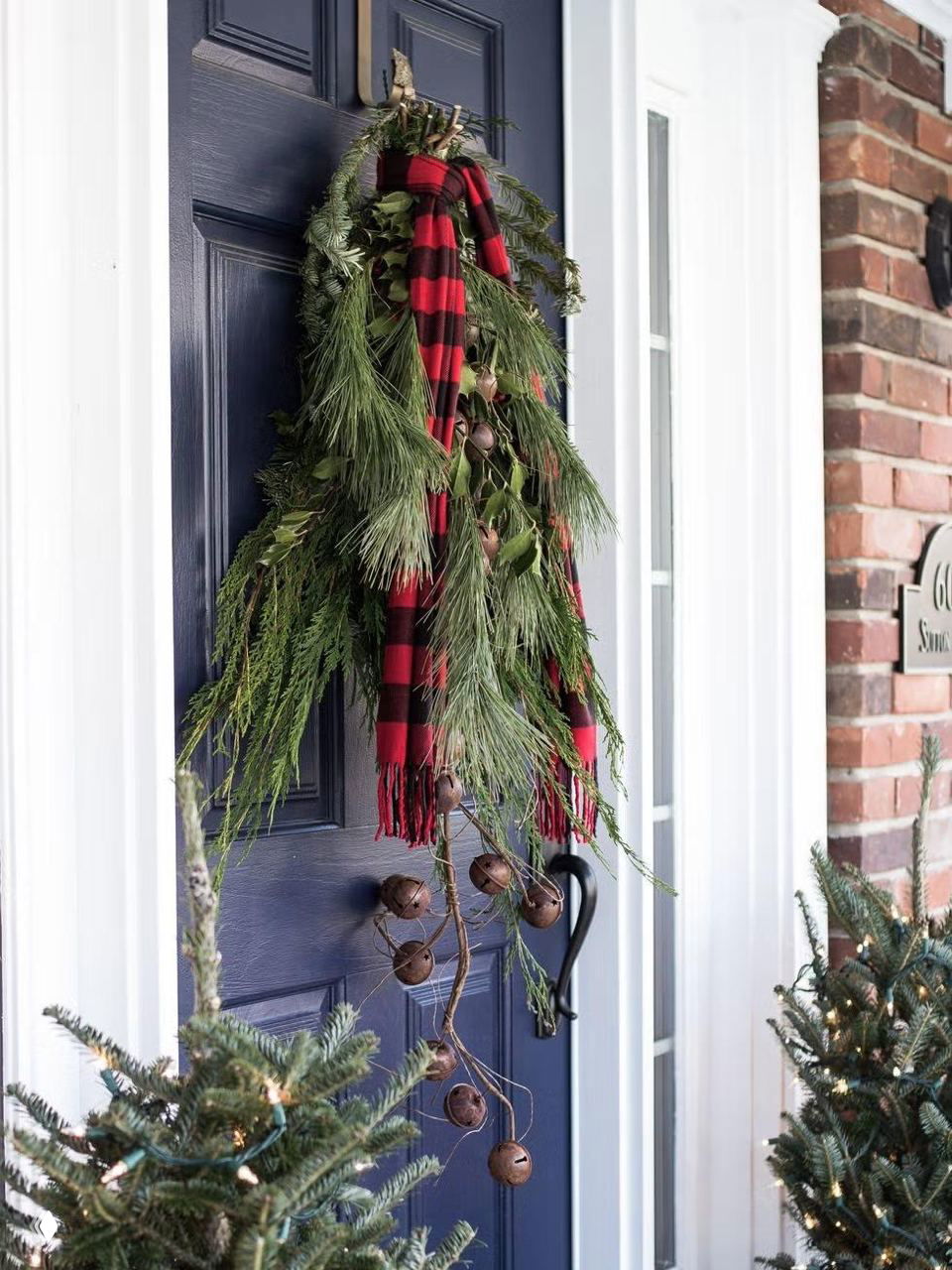 Темно‑синяя дверь украшена вертикальным подвесом из хвои с бубенцами и клетчатым шарфом — простой деревенский стиль для входа в дом в холодный сезон.