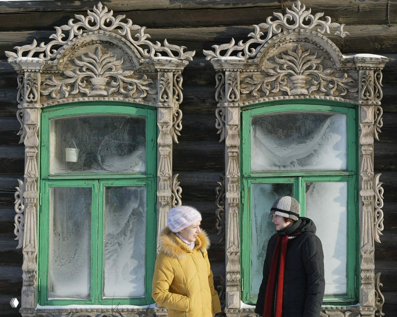 Два человека разговаривают у старого деревянного дома с зелёными рамами окон и богато резными наличниками — фрагмент традиционного томского зодчества.
