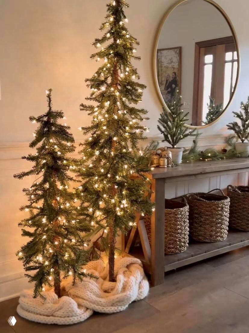 Тонкие подсвеченные ёлки у консоли с круглым зеркалом, тёплый свет гирлянд подчёркивает рельеф ветвей и создаёт уютную праздничную сцену.