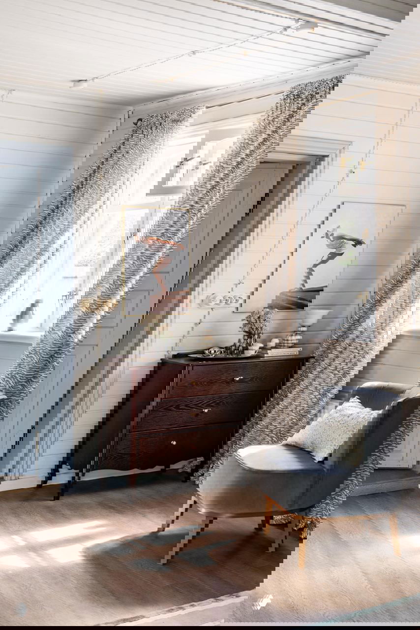 Уютный интерьерный уголок: тёмный комод, серое кресло, длинные шторы и солнечный свет на деревянном полу в дачном доме.