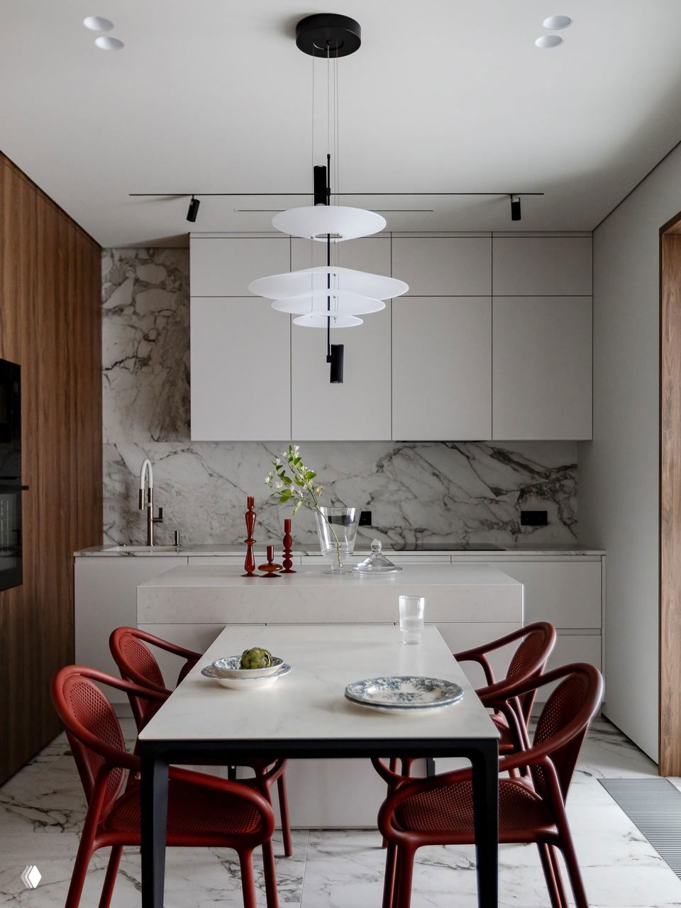 Обеденная зона с тонкой столешницей и красными стульями, на заднем плане мраморный фартук и минималистичные белые кухонные фасады — акцент на формах и текстурах.