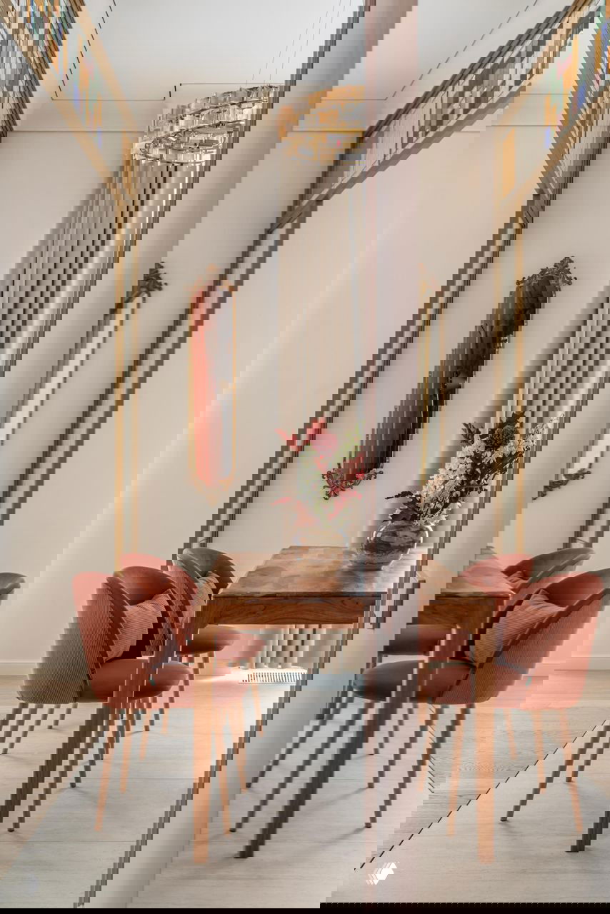 Отражённый снимок столовой: вид через зеркальную/стеклянную перегородку, деревянный стол, терракотовые стулья и декоративное зеркало на стене.