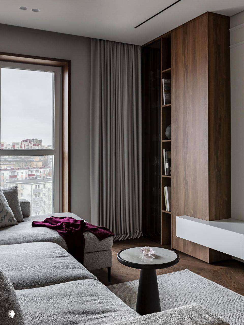 Гостиная с большим угловым диваном нейтрального цвета, деревянными панелями и видом из окна на городской пейзаж; текстуры ткани и дерева гармонируют с лепниной.