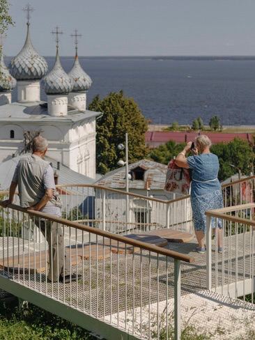 Пешеходы поднимаются по металлической лестнице с перилами; вокруг видны исторические крыши и зелень города Юрьевца.