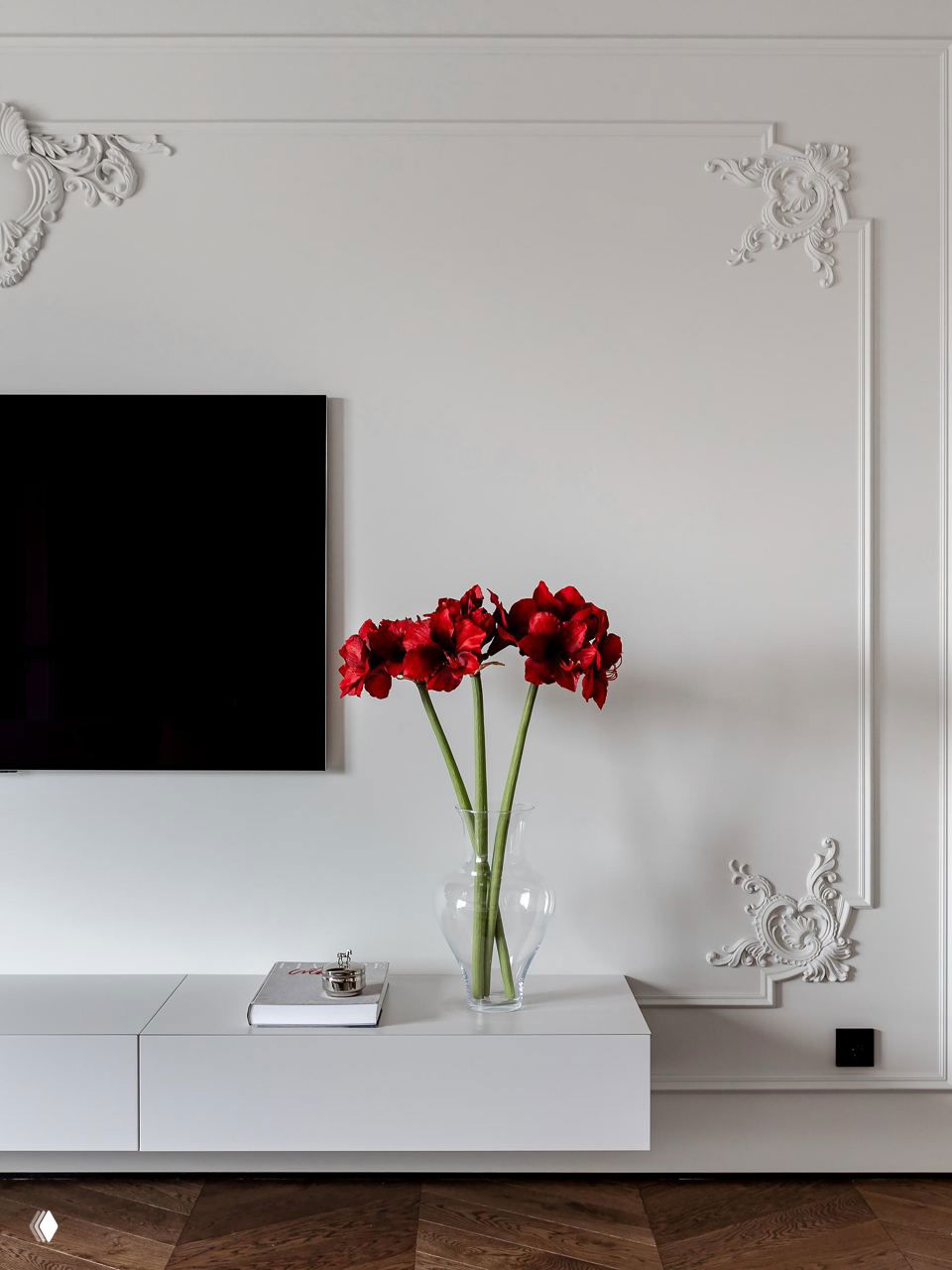 Декоративная лепнина на светлой стене рядом с телевизором, вазой с ярко-красными цветами — контраст классики и современного минимализма в деталях интерьера.