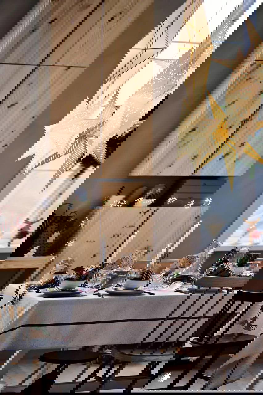 Общий план столовой: белая скатерть, свечи и парящие бумажные звезды‑фонари над празднично украшенным столом в светлом интерьере