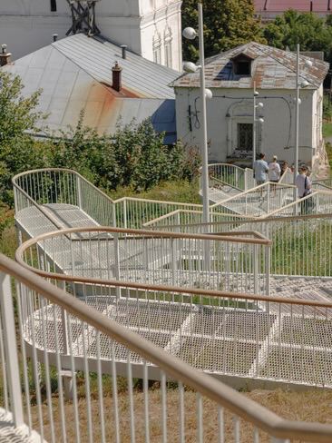 Крупный план изогнутой белой ограждённой лестницы и переходов; сетчатые ступени и деревянные поручни в летнем освещении.