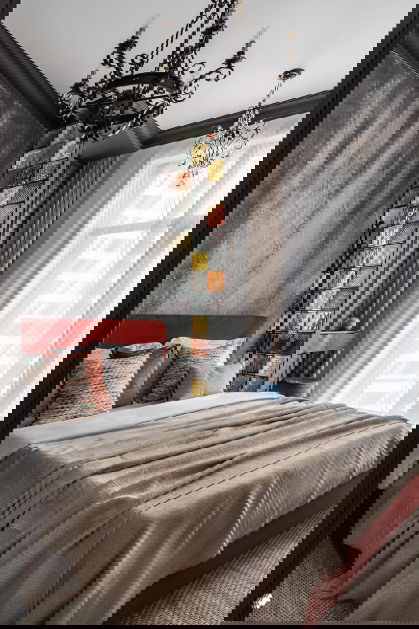 Спальня с акцентной стеной из цветных стеклоблоков и высокими окнами: кровать с терракотовым изголовьем, бархатные подушки и декоративные люстры.