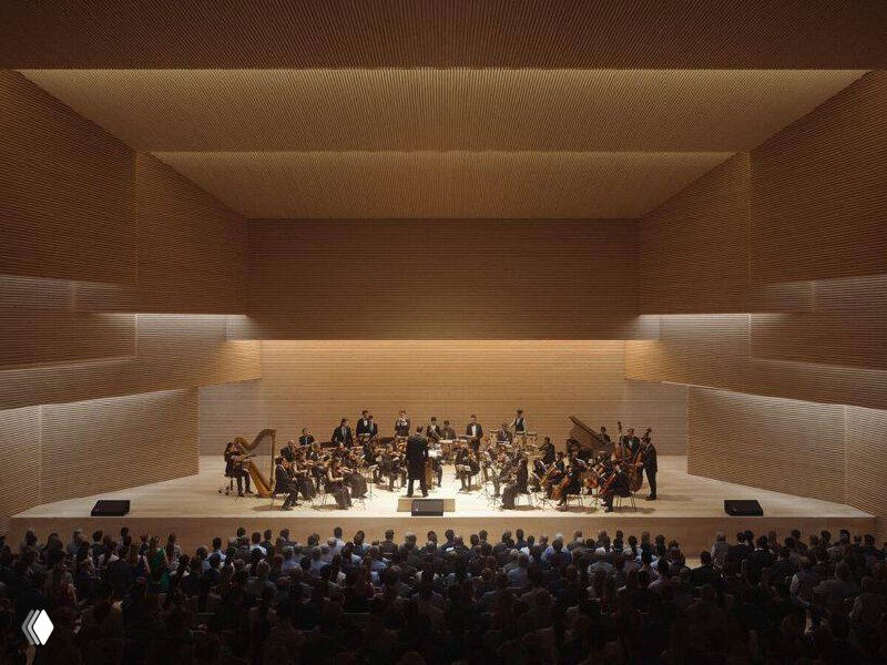 Большой зал или аудитория с трибунами на фоне тёмной отделки: зрители и сцена на большом пространстве, ощущение масштабности.