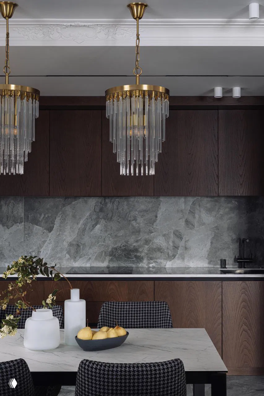 Кухонный фартук из серого камня, тёмные деревянные шкафы до потолка и декоративные подвесные люстры со стеклянными трубками вблизи рабочей зоны.