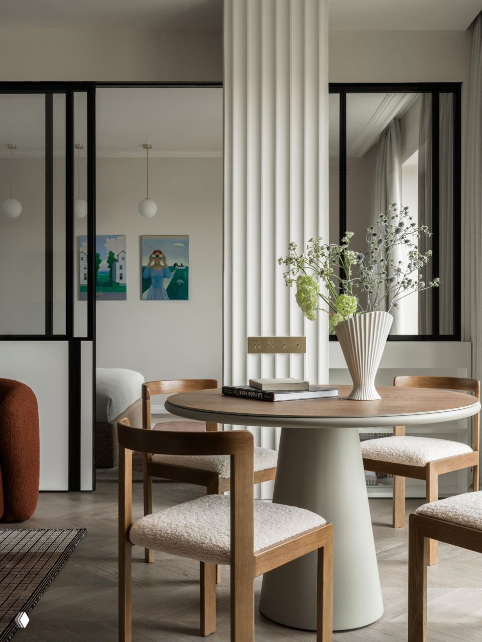 Обеденная зона с круглым столом и деревянными стульями на фоне вертикальной декоративной колоны и больших окон, светлая спокойная гамма.