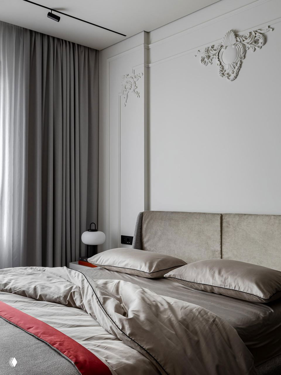 Спальня с красивой декоративной лепниной над изголовьем, мягкие текстуры постельного белья и спокойная нейтральная палитра с небольшими красными акцентами в текстиле.