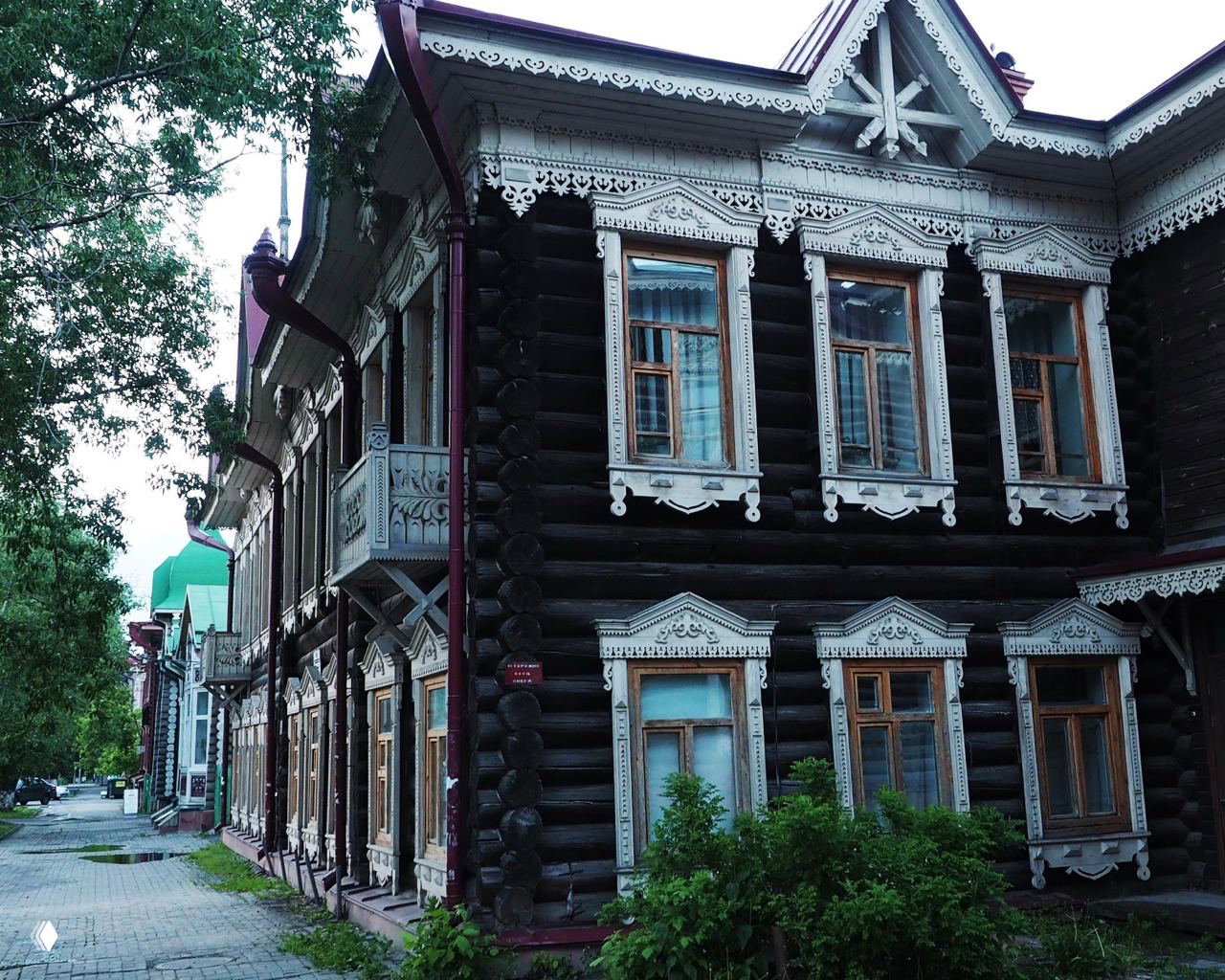 Двухэтажный тёмный бревенчатый дом с белыми резными наличниками и балконами вдоль тихой улицы Томска — типичный образец деревянного зодчества.