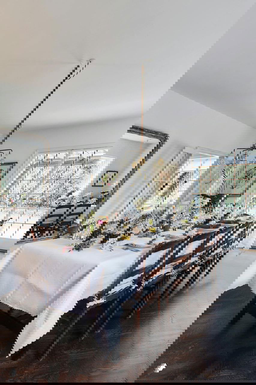 Большой обеденный стол, накрытый белой скатертью, сервировка стеклом и фарфором, высокие окна и хрустальная люстра над столом.