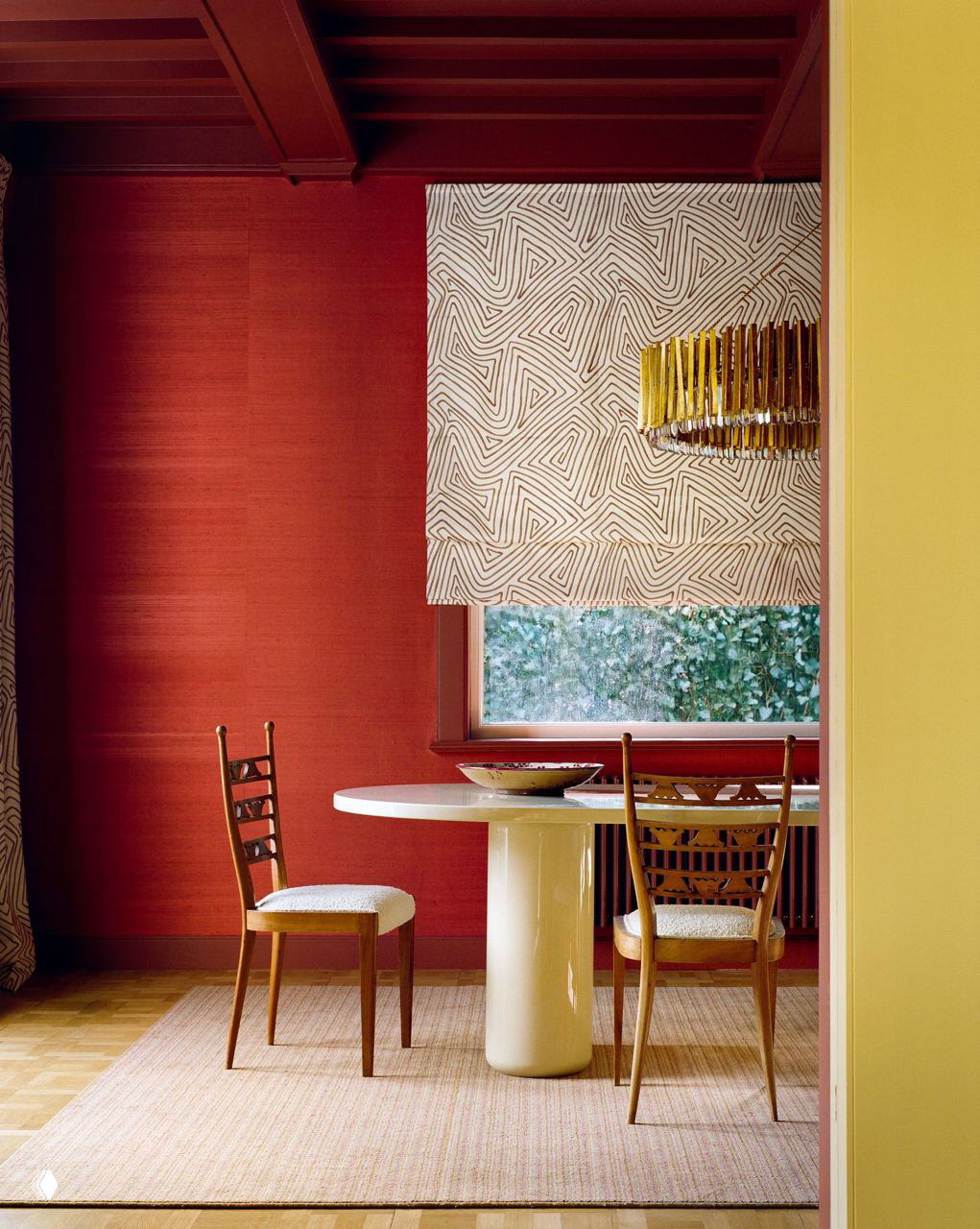 Кадр столовой с круглым белым столом у окна, бордовые стены и текстурные римские шторы на фоне; видна часть солнечной жёлтой стены рядом.