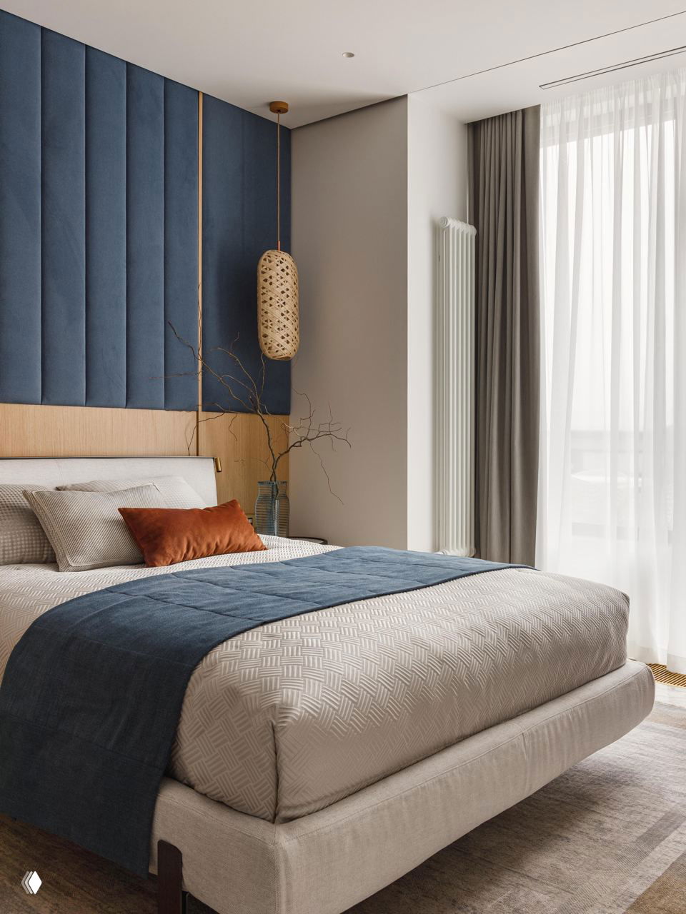 Спальня с высокой обивкой из синего текстиля над изголовьем, светлая постель с синим пледом и оранжевой декоративной подушкой для цветового контраста.