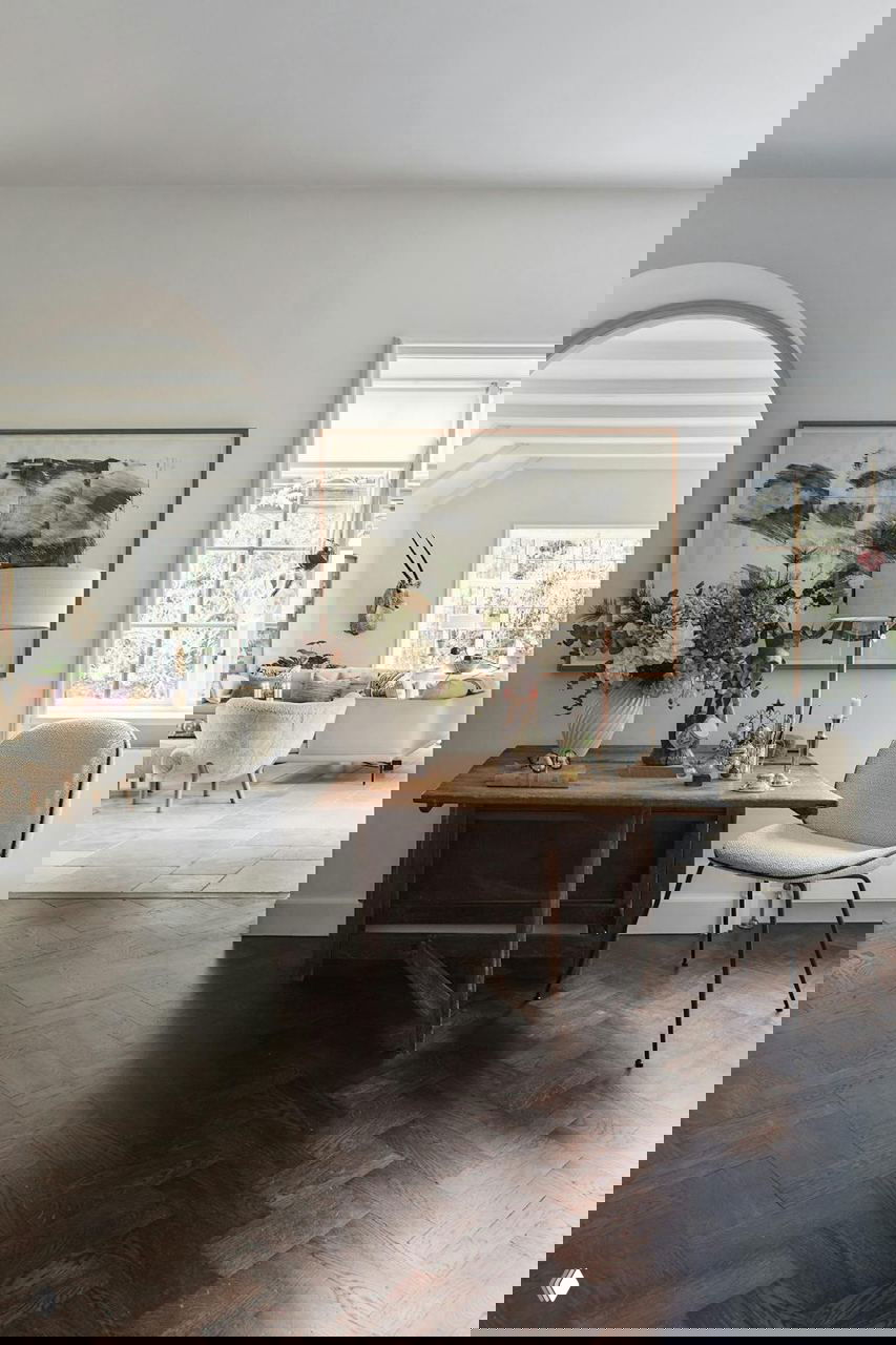 Прихожая через арку: деревянный стол с вазой цветов, декоративные предметы и стул на фоне светлого интерьера и тёмного паркета.